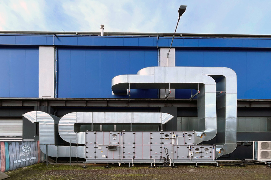 Das Wolf RLT-Gerät KG Top mit geschwungenen Zu- und Fortluft-führenden Kanälen steht im Außenbereich auf dem Boden neben der Halle - es hätte auf dem geschwungenen Hallendach nicht platziert werden können.