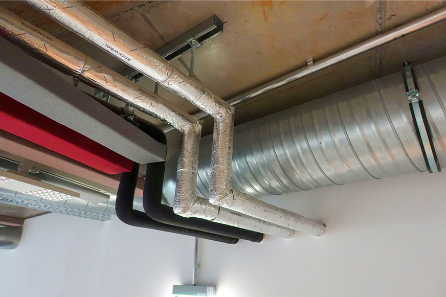 Deckengebundene Leitungen und farbige Schallschutzlamellen. Die Leitungen für Fußbodenheizung und Warmwasser sind gegen Wärmeverlust gedämmt, die Kaltwasser- und Regenwasserleitung gegen Wärmeaufnahme – letztere sind ­zusätzlich gegen Kondenswasserbildung diffusionsdicht ummantelt.