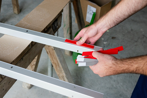 Die Montageplatten werden zusammen mit der Fixierhilfe in die Langlöcher des Wandanschlussprofils gesteckt. Die Klebeschablone wird auf die Montageplatte aufgebracht.