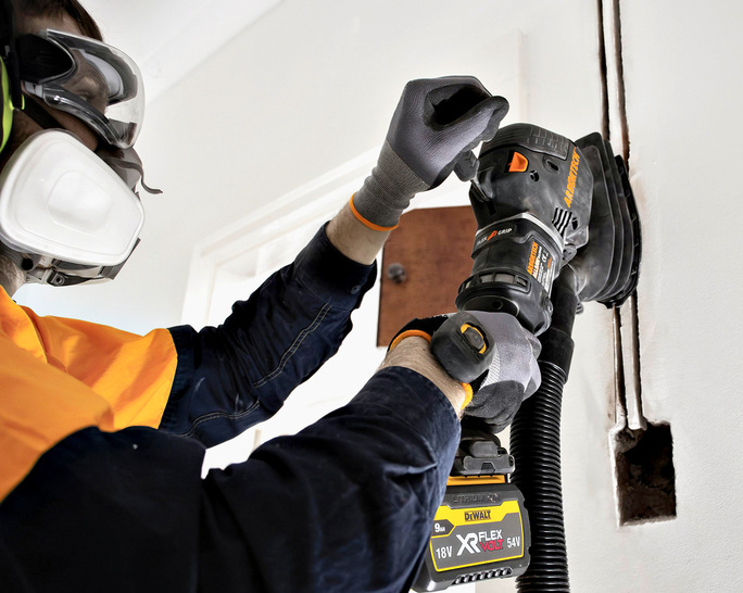 „SHK-Ausbildender trägt Schutzbrille und Gehörschutz, verwendet einen Bandschleifer zur glatten Oberflächenbearbeitung einer Trockenbauwand in einem Heizungsraum. - © Bild: Arbortech