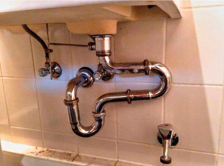 "Installierte Chrom-Trinkwasserleitungen unter einem Waschbecken vor einer weiß gefliesten Wand, exemplarisch für moderne Sanitärinstallationen im SHK-Handwerk. - © Bild: Döhring