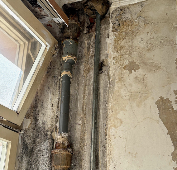 Offenes Fenster neben einer fleckigen, rissigen und schimmligen Wand mit freiliegenden Rohren in einem baufälligen Raum. - © Bild: Wolfgang Jörger