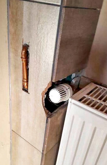 Ein Heizkörperventil ragt unbeholfen durch ein gebrochenes Loch in der gefliesten Wand neben einem Heizkörper heraus.