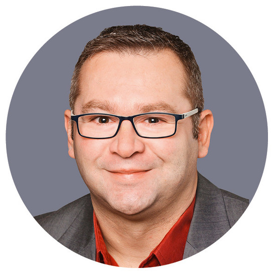 Lächelnder Mann mit kurzen braunen Haaren, Brille und einem grauen Blazer über einem roten Hemd, auf grauem Hintergrund.