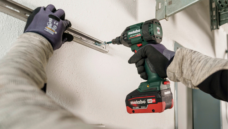 Person, die mit einem Akkubohrer eine Metallschiene an eine weiße Wand schraubt, trägt Handschuhe und Arbeitskleidung. - © Bild: Metabo