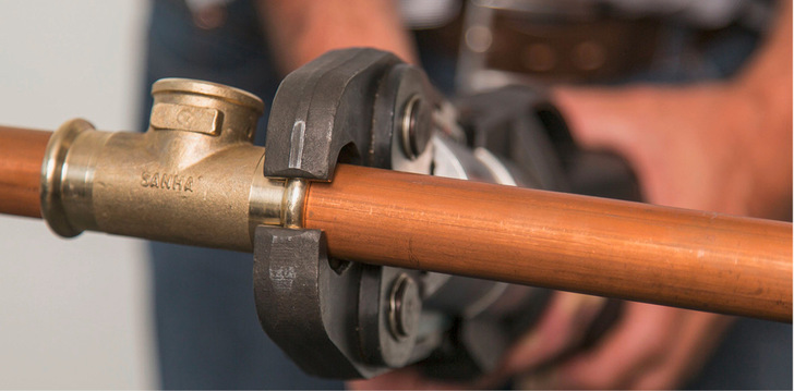 Nahaufnahme eines Werkzeugs, mit dem ein Fitting auf ein Kupferrohr gepresst wird, gehalten von einer Person im Hintergrund. - © Bild: Sanha