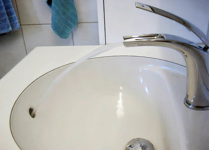 In einem Badezimmer mit blauen Handtüchern sprudelt ein Wasserhahn außerhalb des Waschbeckens, das Becken fehlt.