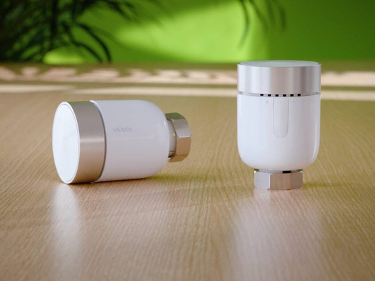 Alt-Text: „Zwei weiße, zylindrische Smart-Home-Geräte mit metallischen Akzenten auf einem Holztisch, die innovative Lösungen für die Steuerung von Heizungs-, Lüftungs- und Klimatechnik in vernetzten Gebäuden demonstrieren; grüner Hintergrund. - © Vilisto