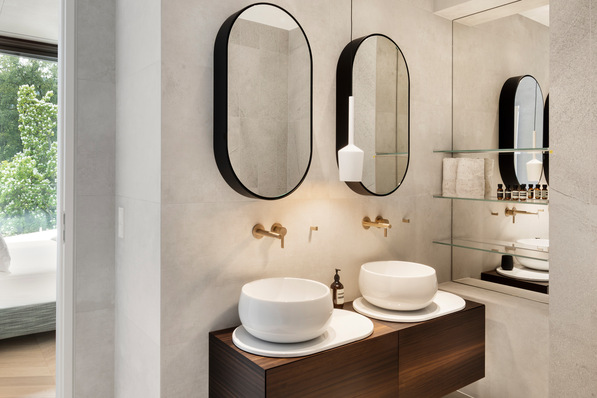 Alt-Text: „Modernes Badezimmer mit doppeltem Waschbecken, ovalen Spiegeln und goldenen Armaturen. Große Fensterflächen sorgen für natürliche Beleuchtung und tragen durch Wärmeisolierung zur Energieeffizienz bei. Passend für zeitgemäße Lösungen in der Sanitärinstallation. - © Ken Schluchtmann, diephotodesigner.de