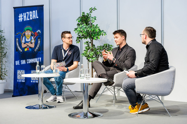 „Drei Fachleute diskutieren in modernen Sesseln bei einem Podiumsevent über neue Trends in der Gebäudetechnik, im Hintergrund ein Banner zur Sanitär-, Heizungs- und Lüftungstechnik sowie eine Zimmerpflanze. - © Messe Essen