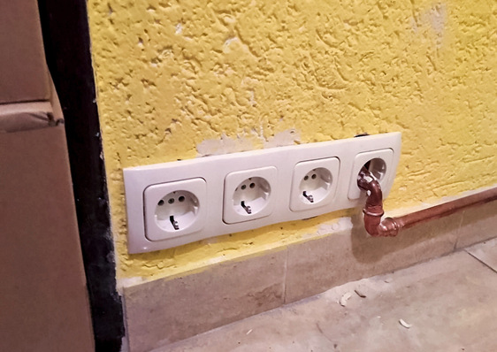 Vier weiße Steckdosen an einer gelben Wand, die teilweise durch ein Kupferrohr verdeckt ist.